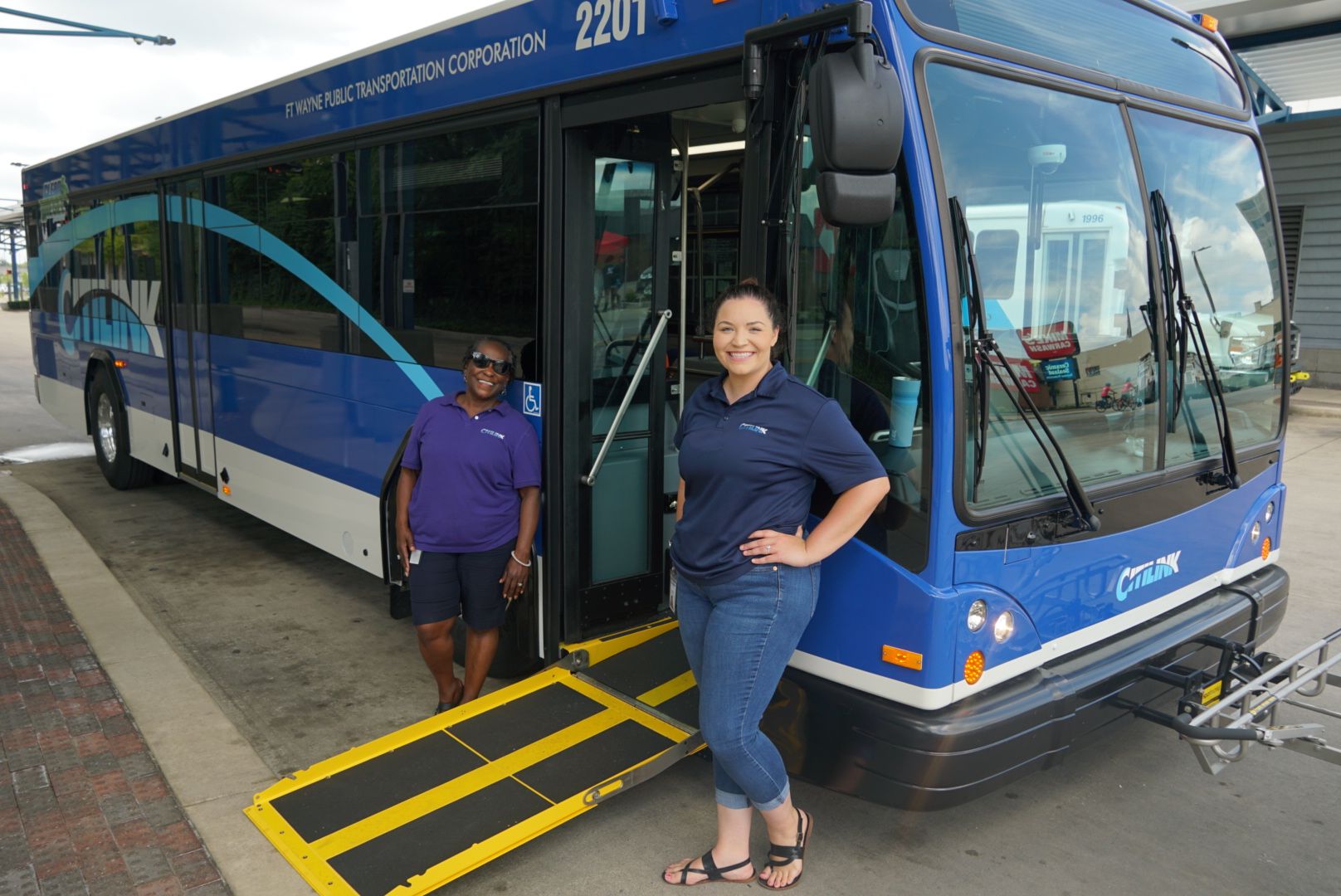 Two women stand next to a wheelchair ramp leading to the open door of a Citilink bus