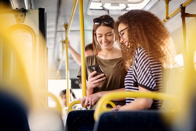 Two young smiling women look at a phone while standing on the bus